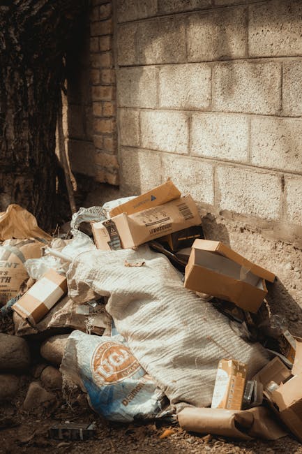 A pile of discarded rubbish next to a brick and concrete wall, consisting of cardboard boxes, plastic packaging, and paper waste, some of which are crushed or flattened. The cardboard boxes vary in size and are mostly brown with printed labels and barcodes visible on their surfaces. A textured white sack or bag is partially visible within the clutter, along with smaller pieces of packaging material. The rubbish is loosely piled on the ground, which appears to be dirt or gravel, with a large tree trunk partly visible on the left side of the image. The background shows the wall made of large, light-colored concrete blocks, with a darker brick section at the corner, suggesting an outdoor or semi-enclosed space used for waste storage or temporary rubbish accumulation. The scene reflects an area that could be prepared for domestic or commercial rubbish removal, aligning with the services offered by Rubbish Removal Belsize Park for efficient private waste disposal or on-site clearance.