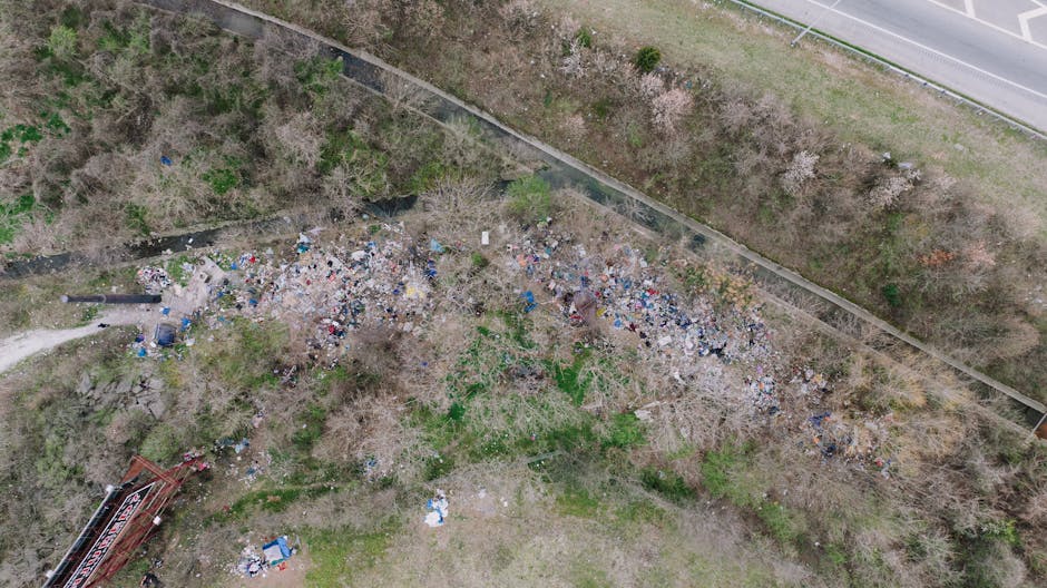 An aerial view of a large area of unmanaged outdoor rubbish and debris scattered across a patch of land adjacent to a footpath and a low fence. The waste consists of various materials, including plastic bags, cardboard, and other refuse, with some items appearing crumpled or torn. Surrounding the debris are leafless trees and sparse green vegetation, indicating early spring or late autumn. In the background, a paved pathway runs parallel to the fence, while a railway track runs along the top edge of the image, separated from the rubbish site by a strip of grass and bushes. The scene appears to be a neglected or unmanaged waste site and highlights the importance of proper rubbish disposal and collection, such as those provided by Rubbish Removal Belsize Park, to prevent litter accumulation and environmental degradation.