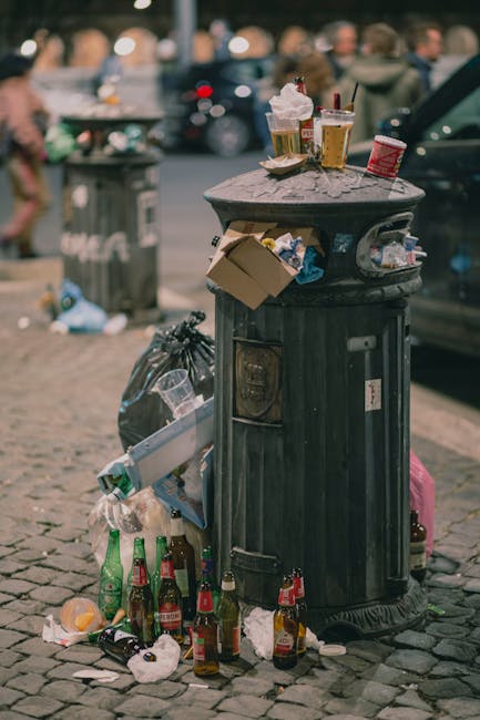 A pile of discarded rubbish next to a brick and concrete wall, consisting of cardboard boxes, plastic packaging, and paper waste, some of which are crushed or flattened. The cardboard boxes vary in size and are mostly brown with printed labels and barcodes visible on their surfaces. A textured white sack or bag is partially visible within the clutter, along with smaller pieces of packaging material. The rubbish is loosely piled on the ground, which appears to be dirt or gravel, with a large tree trunk partly visible on the left side of the image. The background shows the wall made of large, light-colored concrete blocks, with a darker brick section at the corner, suggesting an outdoor or semi-enclosed space used for waste storage or temporary rubbish accumulation. The scene reflects an area that could be prepared for domestic or commercial rubbish removal, aligning with the services offered by Rubbish Removal Belsize Park for efficient private waste disposal or on-site clearance.