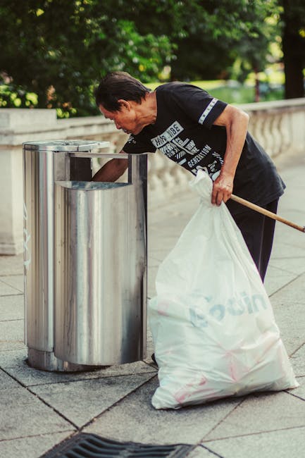 A middle-aged man with dark hair, wearing a black T-shirt with white graphic text on the front, is seen disposing of rubbish into a silver metallic waste receptacle situated on a sidewalk. He is bending slightly forward, holding a long-handled tool in his right hand, which is used to position a large, partially filled white plastic garbage bag in front of the open-top bin. The man’s left hand stabilizes the bag, which appears to contain miscellaneous waste. The background features lush green foliage and a stone balustrade, indicating he is outdoors in a park or urban green space. The pavement is composed of square concrete slabs, and the scene is illuminated by natural daylight, casting soft shadows. The image relates to independent waste collection services, such as those offered by Rubbish Removal Belsize Park, emphasizing proper waste disposal in communal outdoor areas while highlighting the process of rubbish removal in a public setting.