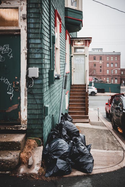 A small urban property with a teal wooden exterior featuring horizontal siding and red-trimmed windows. The building has a set of wooden stairs leading up to a white front door, partially visible on the right side of the image. Near the base of the building, on a concrete sidewalk, there are multiple tightly packed black rubbish bags that appear to be filled with waste, placed beside a patch of brown, dried grass. To the left, part of a graffiti-covered metal door or shutter is visible, showing signs of weathering and rust. In the background, a parking lot with a few cars, including a black vehicle parked on the right, extends toward a large brick industrial building or warehouse in the distance, under an overcast sky. The scene suggests a setting where private waste collection or disposal services, such as those offered by Rubbish Removal Belsize Park, may be involved in clearing accumulated rubbish from urban properties, emphasizing the practical need for rubbish removal and waste management solutions in city environments.