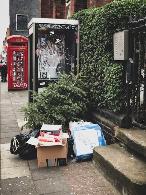 A collection of discarded household items and waste materials placed on a city sidewalk in front of a small, partially covered kiosk and a green hedge. The waste includes a partially flattened cardboard box, a white plastic bag, and a blue and white cardboard box, all stacked among a few loose items. Nearby, a small potted evergreen tree lies on its side, contributing to the cluttered appearance. Behind the waste, a black metal fence with a hedge growing over it separates the sidewalk from an adjacent property. The scene also features a traditional red British telephone booth further along the pavement, and a nearby grey concrete step leading up to an entrance, indicating an urban environment. This setting suggests a situation where private waste collection or rubbish removal services, such as those provided by Rubbish Removal Belsize Park, might be involved in clearing such rubbish from public or private spaces in the area.