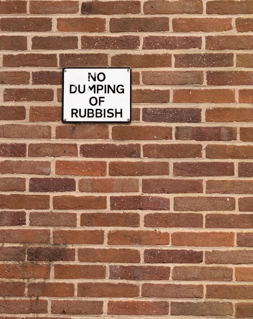 A rectangular metal sign attached to a red brick wall, displaying the message 'NO DUMPING OF RUBBISH' in black uppercase letters on a white background. The sign is positioned near the upper left section of the brick surface, which features evenly arranged bricks in shades of reddish-brown and dark brown, with light mortar in between. The brick wall extends vertically and horizontally, with no other objects or markings visible nearby. The lighting appears natural, casting minimal shadows on the wall, indicating an outdoor setting consistent with urban or residential environments where rubbish removal services like those of Rubbish Removal Belsize Park are relevant.