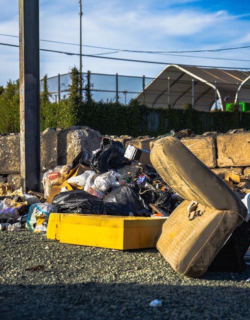 A pile of mixed rubbish and waste materials situated on the ground next to a stone wall, with black garbage bags, a yellow plastic tray, and an old, worn car seat with fabric upholstery in beige tones. The waste includes plastic packaging, papers, and other debris, scattered on a gravel surface. In the background, there is a metal fence with a green hedge behind it, and a large white tent-like structure with metal supports, set against a partly cloudy sky with blue and white hues. A utility pole with wires is visible on the left side of the image, indicating an urban or semi-urban environment. The scene appears to be an instance of informal rubbish accumulation in an outdoor area, possibly during an on-site waste clearance or disposal process managed by a waste removal service such as Rubbish Removal Belsize Park, situated near Haverstock Hill NW3.